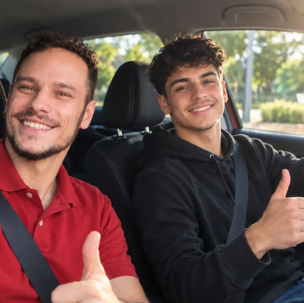 Fahrlehrer und Fahrschüler im Auto, beide lachen und zeigen den Daumen nach oben